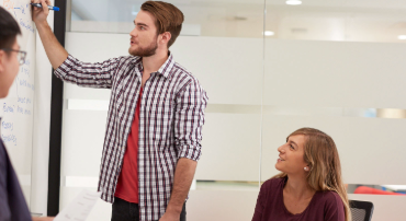 Three students in a glass-walled study room, with one man writing on a whiteboard while a seated woman looks up at him and listens attentively, the other man is standing to listen.