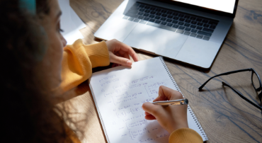 A top-down shot of a student writing maths equations on a notebook with an open laptop computer