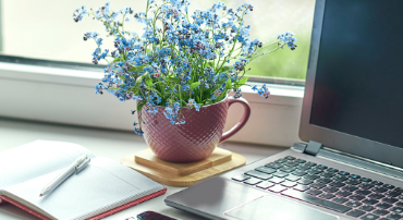 A potted flowering plant in a pink mug sits beside a laptop and notebook.