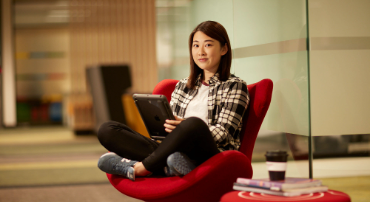 Student sits on a bright red chair, cross-legged and holding a large handheld tablet.